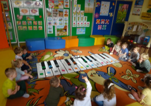 Dzieci siedzą wokół rozłożonego na dywanie instrumentu Kolorpiano.